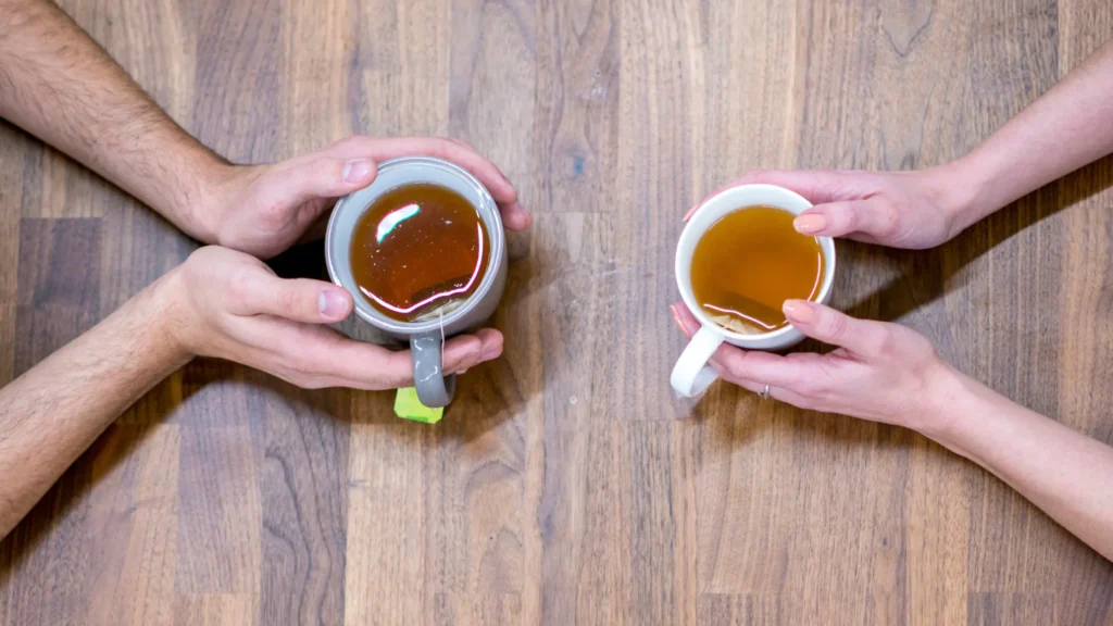 Top-down view of two people holding ceramic mugs of freshly brewed tea over a wooden table, sharing a conversation.